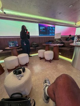 A first-person perspective from a seat showing someone's legs/feet in sneakers and a black bag, looking toward a lounge or event space with green LED lighting, stools, tables with water bottles, and a person standing by monitors in the background.