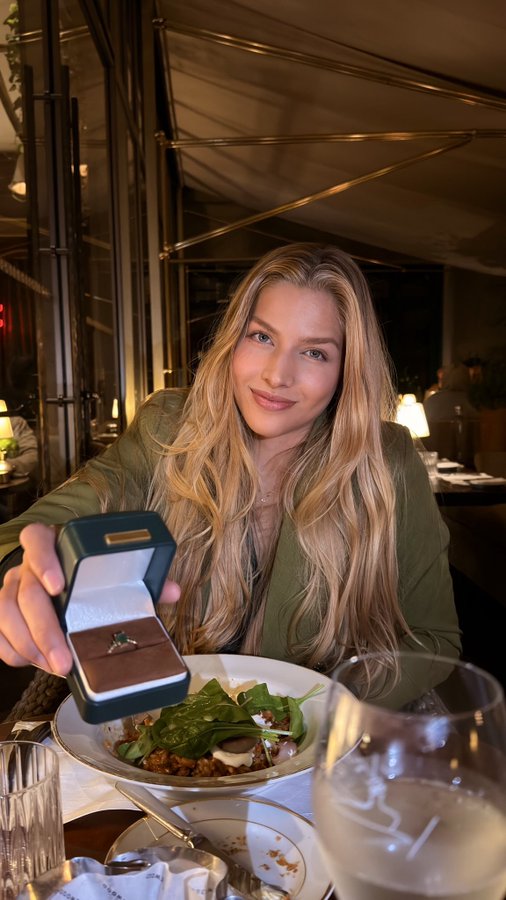 A blonde woman smiles as she presents an open green ring box containing a ring, seated at an outdoor restaurant table with a salad in front of her.
