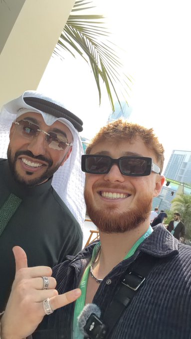 Two men in sunglasses smile and flash a shaka at the camera outdoors; one wears a lanyard marked “VIP.” “GM” greets followers with a bright, upbeat selfie.