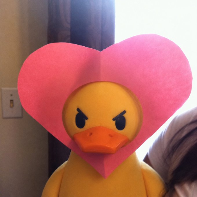 A yellow rubber duck with an angry expression wears a large pink paper heart around its head in a dimly lit room.