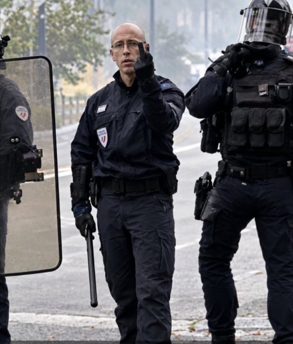 A photograph of a bald police officer in uniform holding a baton, flanked by two riot police officers in full tactical gear with shields.