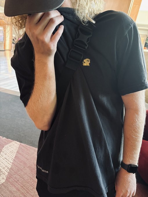 A person wearing a black t-shirt with a small yellow octopus logo, a black cap, and a crossbody bag, covering their face with their hand.