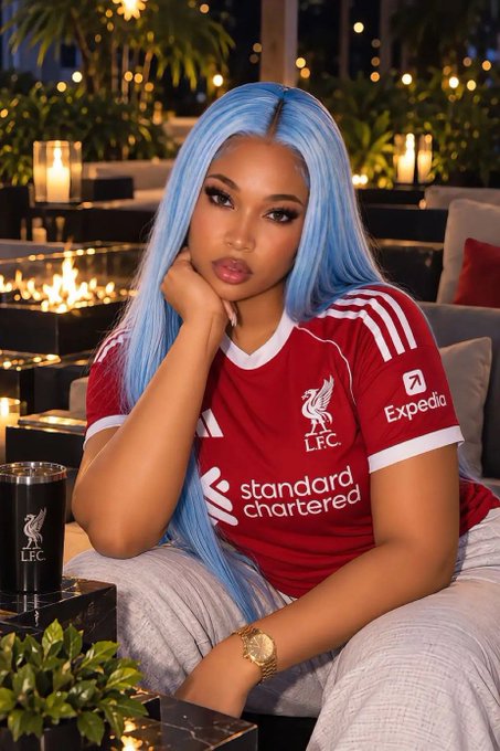 A young woman with long blue hair wearing a red Liverpool FC jersey featuring the LFC crest, Standard Chartered sponsor, and Expedia logo, posing in a candlelit outdoor lounge setting with an LFC-branded cup nearby.