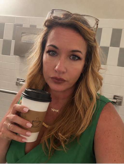 A selfie of a woman with blonde wavy hair wearing a green top, holding a takeaway coffee cup in a tiled restroom, with sunglasses perched on her head and a gold "mama" necklace visible.