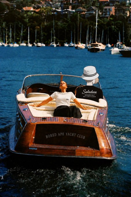 Luxury lifestyle photo of a person lounging on the back of a classic wooden boat named 'Bored Ape Yacht Club' with a 'Saladein' advertisement, cruising on blue harbor waters.