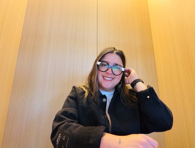 A young woman with long hair and black-rimmed glasses smiling at the camera while sitting against a wood-paneled wall.