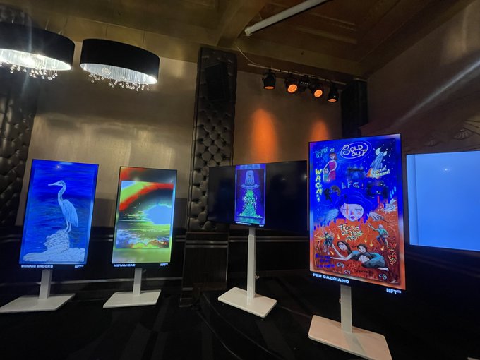 A dimly lit gallery space displays several digital screens showcasing colorful, artistic NFT artworks.