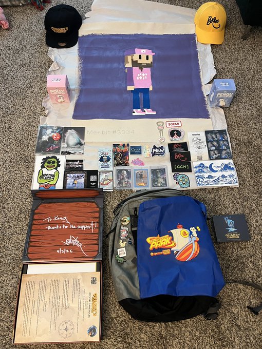 A collection of merchandise laid out on carpet, including two caps (black and yellow with 'BMC' text), a blue pixel-art print of a character labeled 'weebit #3034', numerous stickers and patches, a backpack featuring 'Flash' branding, and a wooden sign reading 'To kdot thanks for the support! -Abyss'.