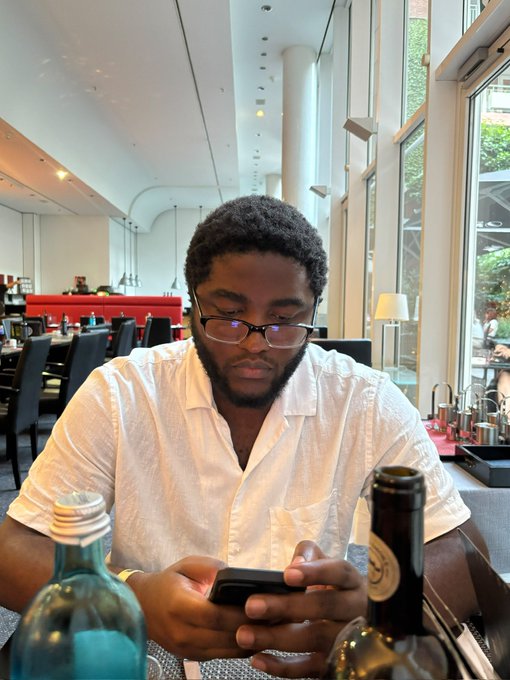 A young man with a beard and glasses wears a white shirt while looking down at his phone in what appears to be a restaurant or hotel lounge, with a water bottle and beer bottle in the foreground. The image aligns with the post's description of a "crypto degen" or "Web3 nerd" engrossed in his phone, possibly checking on trades or airdrops, fitting the "CEO of too many unfinished" projects vibe.