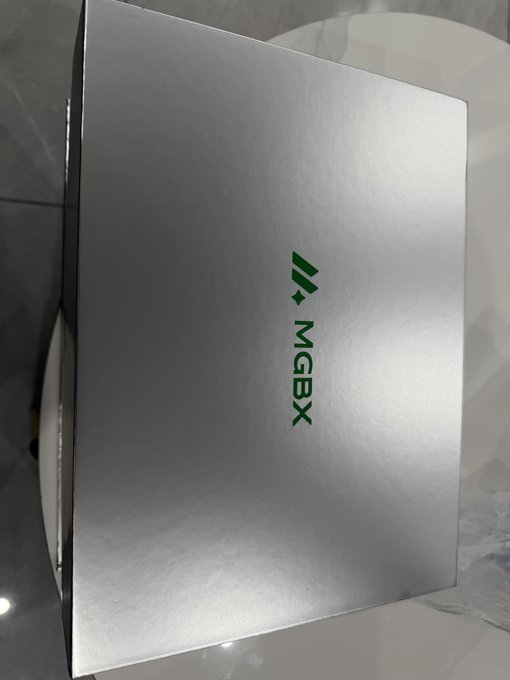A silver cardboard box with the green 'MGBX' logo printed on its top surface, resting on a glossy surface.