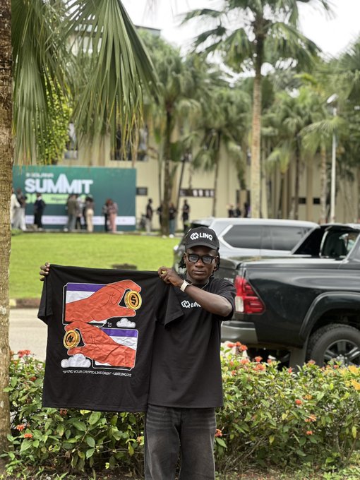 A young man wearing a black cap, glasses, and a t-shirt stands outdoors holding up a black t-shirt with an orange design, with a 'SUMMIT' banner visible in the background.