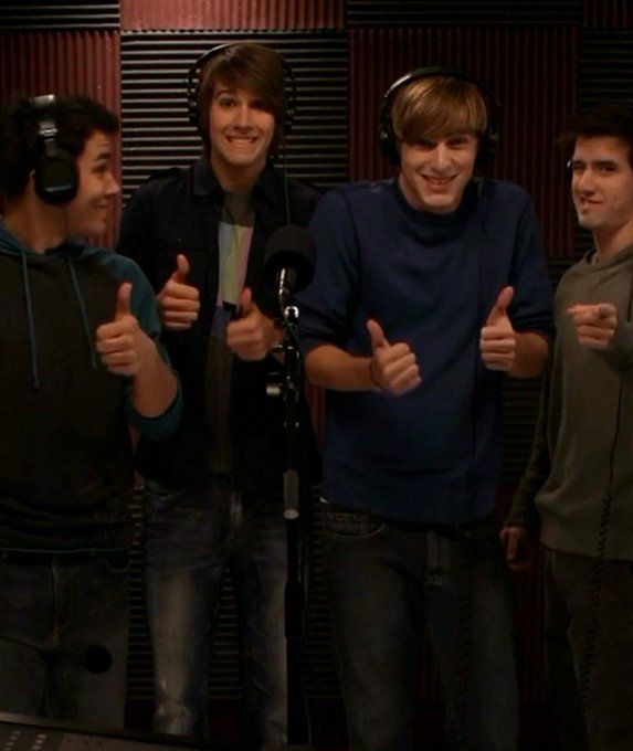 Four young men wearing headphones stand in a recording studio, smiling and giving thumbs-up gestures in front of a microphone.