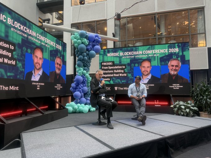The image shows a stage with three large screens displaying images of men sitting, possibly participating in a panel discussion or interview. The screens are positioned at different angles, creating an interesting visual effect as they capture the attention of the audience. In front of these screens, there is a man and a woman seated on chairs, likely engaged in conversation about gemstones. 

The stage also features two potted plants placed near the seating area, adding a touch of greenery to the scene. The backdrop includes a building with a large window, providing an urban setting for this event. A balloon arch is present at one end of the stage, further enhancing the visual appeal and atmosphere of the gathering.