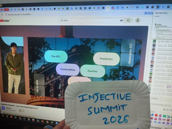 A computer screen shows an Injective Summit live stream from Washington D.C. with category tags (Pre-IPO, Prediction, Commodities, Equities), while someone holds up a paper sign reading 'INJECTIVE SUMMIT 2026'.