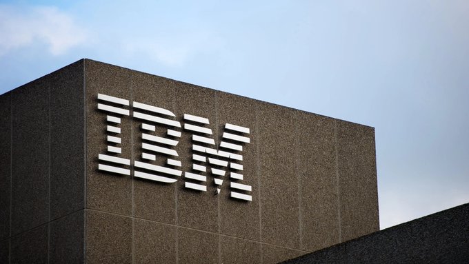 An IBM corporate building sign featuring the iconic striped IBM logo on a brown granite facade against a blue sky.