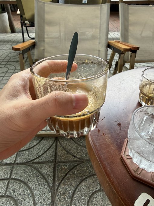 A hand holds a glass of Vietnamese iced coffee (cà phê pha phin) with a black spoon at an outdoor café, accompanied by a glass of water on a wooden table. The image captures the casual café moment referenced in the post's playful "Phê pháo gì chưa" (a pun on coffee "phê" and excitement) morning greeting to friends.