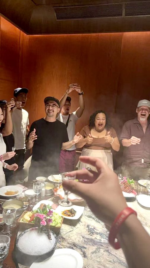 A group of people cheer and laugh around a table filled with food and drinks during a lively gathering.