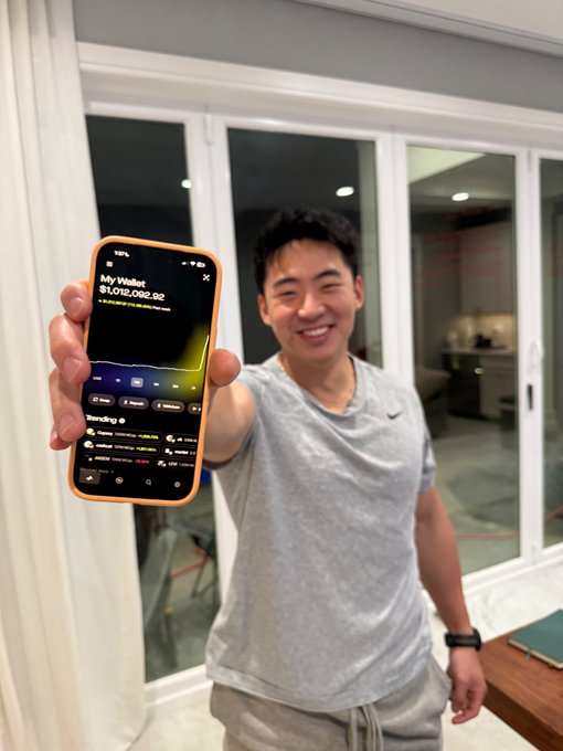 A smiling young man proudly holds up his phone displaying a Robinhood portfolio showing "My Wallets" with a balance of **$1,012,082.92**, with some pending transactions visible at the bottom. The image captures the aspirational moment referenced in the post, showcasing what a successful memecoin portfolio milestone could look like.