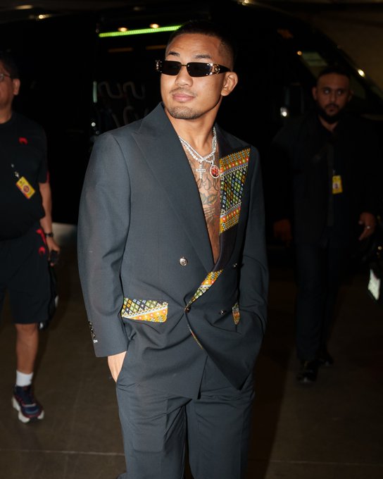 A confident fighter wearing sunglasses and a stylish dark suit with colorful patterned accents poses outside, exuding star power as he arrives for the #CryptoCom #UFC331 main and co-main event.