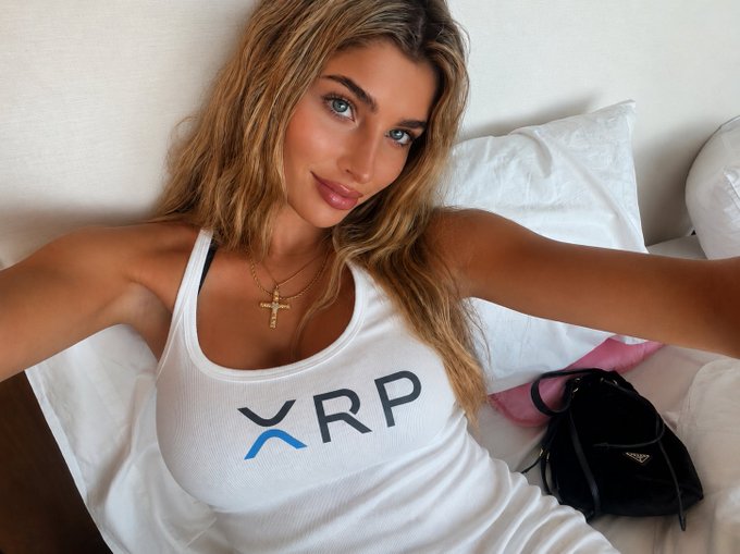A blonde woman in a white tank top featuring the blue 'XRP' crypto logo takes a selfie, wearing a gold cross necklace.