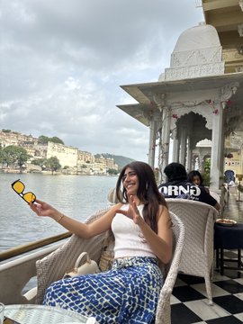 A young woman with long brown hair wearing a white crop top and blue patterned pants sits outdoors at a waterfront cafe, smiling while holding yellow sunglasses, with buildings visible across the lake behind her.