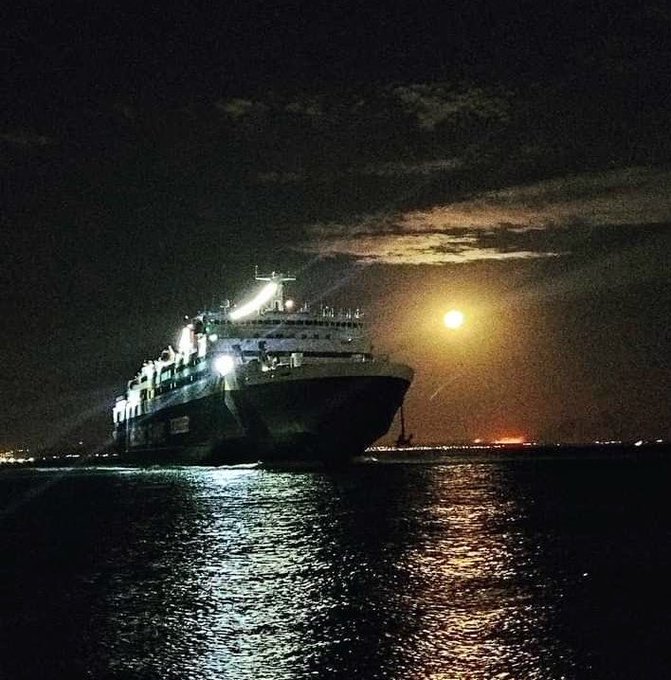 A nighttime photograph of a large illuminated cruise ship or ferry on the water, with the moon glowing brightly in the dark sky above and its reflection shimmering on the water's surface.