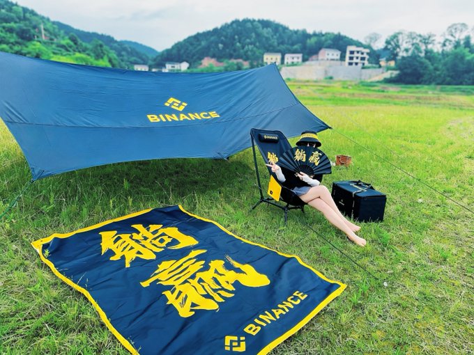 A person relaxes in a chair under a Binance-branded canopy at a scenic outdoor location, with a navy and yellow Binance banner displaying Chinese text in the foreground, tying into Binance's 9th anniversary giveaway promotion featuring Kung Fu Women's Football tickets.