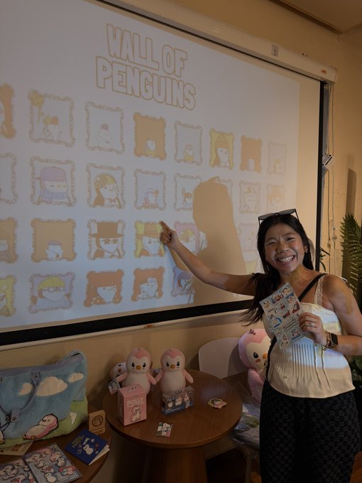 A smiling woman points at a projected 'WALL OF PENGUINS' display featuring various penguin character illustrations, with plush penguin toys and merchandise arranged on a table in front of her at what appears to be a Sanrio event.