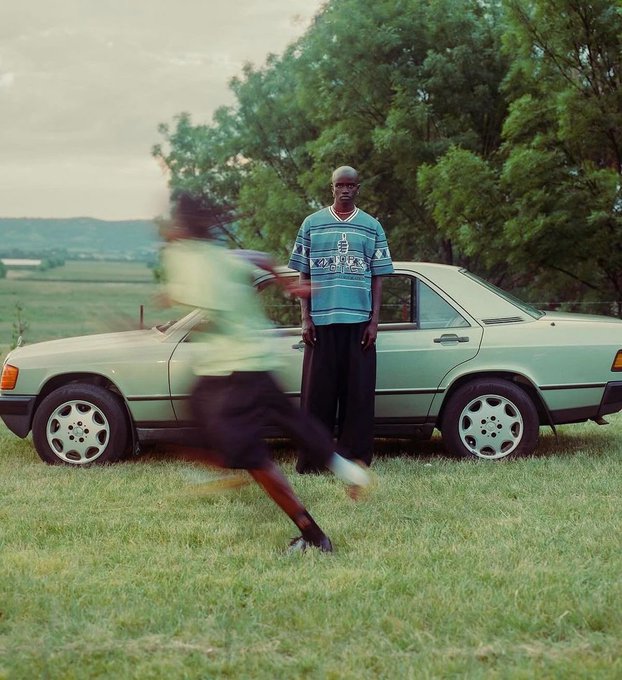 A photo of a man in a blue Kappa shirt standing next to a vintage light green Mercedes-Benz in a grassy field, with another person running past in motion blur.