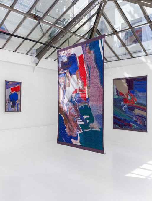 Three large, vibrant textile artworks in shades of blue, red, and purple are displayed in a bright gallery space with a glass ceiling, including a prominent central hanging tapestry that reflects the natural light streaming into the room. The title "Ghost in the Thread" aptly captures the ethereal, woven quality of these abstract fiber art pieces suspended within the airy, sunlit architectural setting.