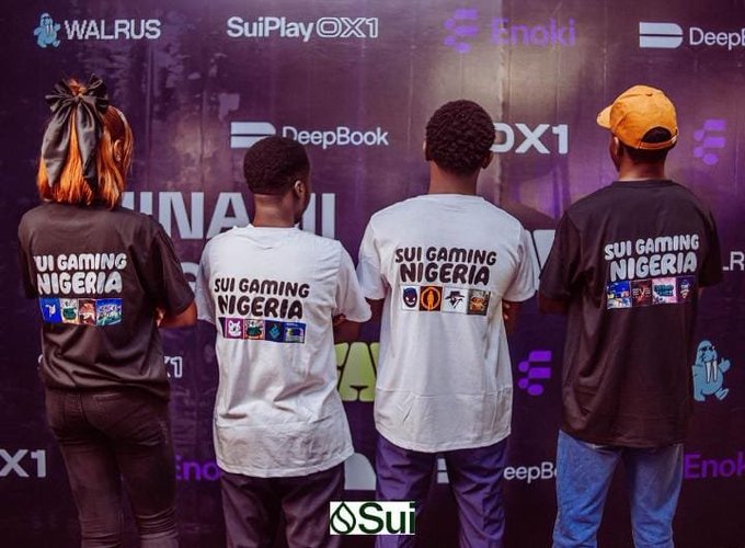 Four people stand with their backs to the camera wearing 'SUI GAMING NIGERIA' t-shirts in front of a backdrop featuring sponsors including Walrus, SuiPlay0X1, Enoki, and DeepBook.