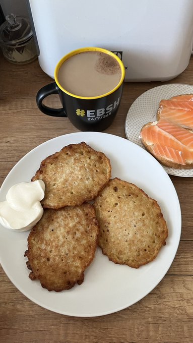 A plate holds three potato pancakes served with a dollop of white cream, accompanied by a mug of coffee labeled 'EBSI' and a slice of salmon on toast.