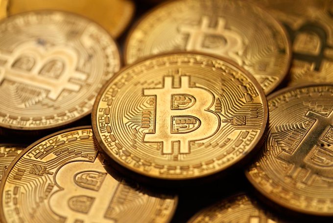 A close-up photograph shows several gold-colored Bitcoin tokens stacked together, prominently featuring the iconic "B" symbol and intricate circuit-like engravings on their surfaces. The image visually represents the article's topic of Bitcoin's price movement, illustrating the cryptocurrency at the center of the news about it hitting a 3-week low amid Strategy's planned $5 billion sale.