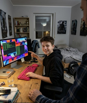 A young man smiles at the camera while sitting at a desk with a pink keyboard and a colorful monitor, with another person partially visible on the right side of the frame.