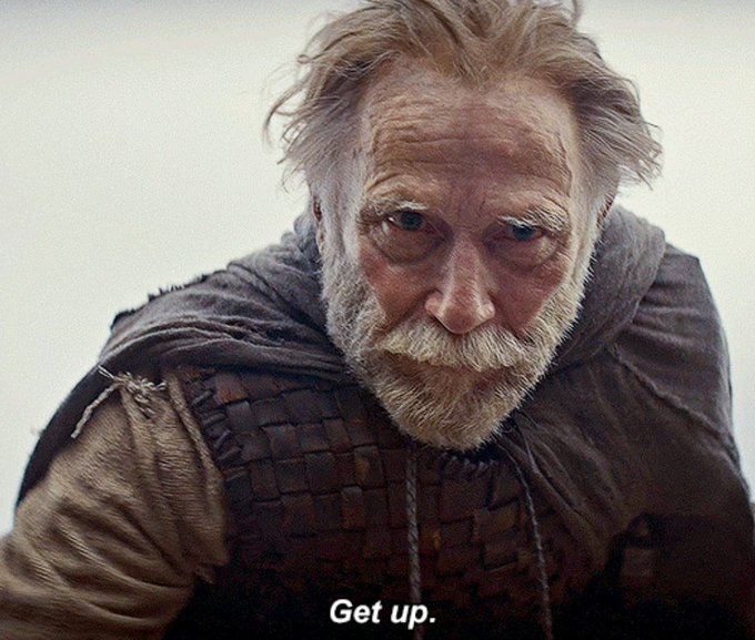 A close-up portrait of an older bearded man in worn, woven medieval-style clothing looking down, with the subtitle 'Get up.'