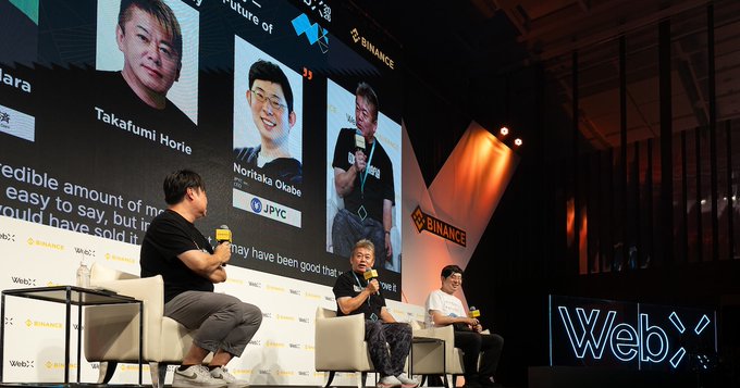 Three panelists sit on stage at the WebX 2026 conference, hosted by Binance and Coinpost, discussing "What About the Japanese Yen in the AI Agent Payment Era?" A large screen behind them displays headshots of featured speakers Takafumi Horie and Noritaka Okabe of JPYC, while the neon "WebX" sign glows to the right of the stage.