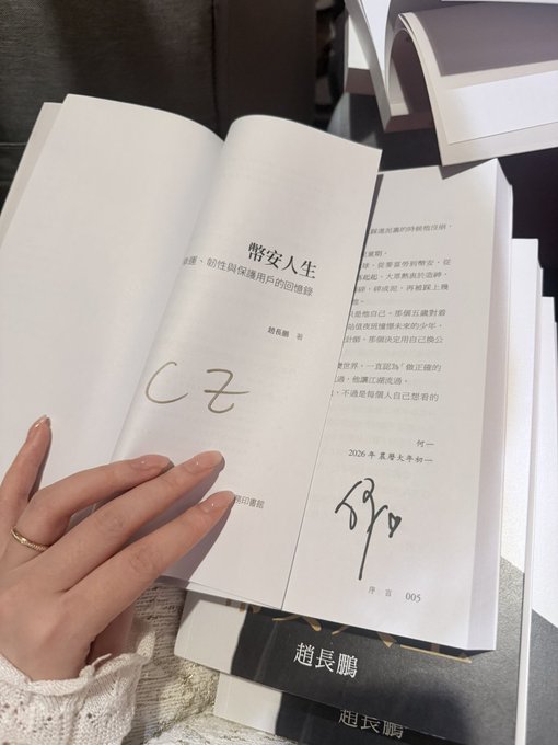 A hand with manicured nails and a gold ring holds open a book titled "幣安人生" (Binance Life), revealing signatures from co-authors Yi Jie and CZ on the title page, with "序言 005" visible at the bottom. The image directly captures the rare dual-signed copy referenced in the post, showing both authors' handwritten signatures alongside the 2026 publication note.