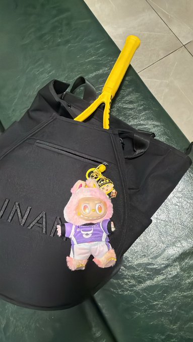 A black duffel bag with 'INAA' text holds a plush keychain of a character in a purple jacket and pink fur hood, with a yellow umbrella tucked inside.