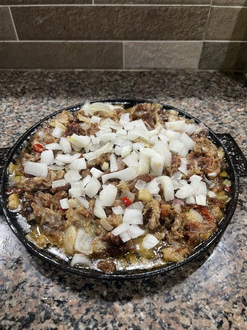 A cast iron skillet filled with a sizzling meat and onion dish topped with diced white onions sits on a granite countertop.