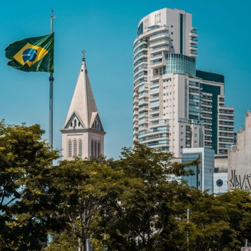A Brazilian flag flies near a historic church and modern skyscraper in Brazil, visually anchoring Tether's $20M investment in Mercado Bitcoin, a local crypto platform expanding tokenization and payments across Latin America.
