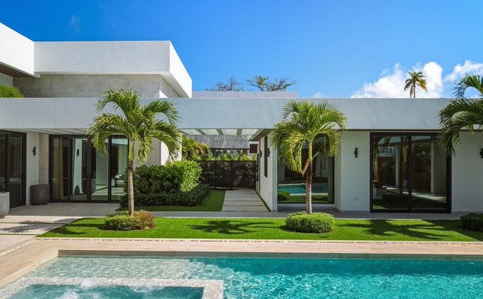 A modern white villa with large glass doors, palm trees in the front yard, and a turquoise swimming pool in the foreground under a blue sky.