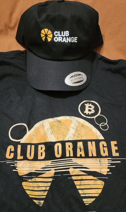 A black baseball cap and matching t-shirt are displayed, both featuring an orange slice logo and the text 'CLUB ORANGE' with a Bitcoin symbol, while the hat's sticker reads 'ADJUSTABLE'.