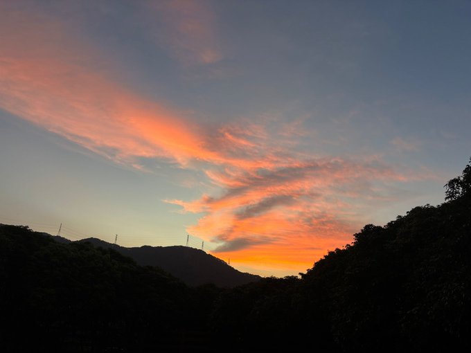 A scenic landscape photograph of a vibrant sunset with pink and orange streaked clouds over a silhouetted mountain range and treeline at dusk.