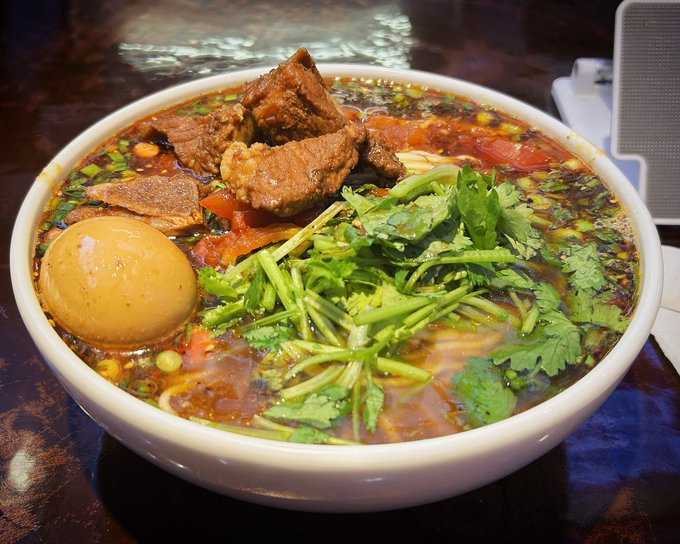 A large white bowl filled with a spicy noodle soup containing beef, a braised egg, and fresh cilantro sits on a dark wooden table.