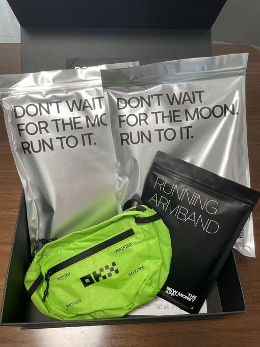 A promotional gift box containing two silver pouches reading 'DON'T WAIT FOR THE MOON. RUN TO IT.', a green running belt, and a black 'RUNNING ARMBAND' package.