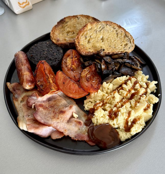 A top-down photograph of a hearty breakfast plate containing bacon, sausage, black pudding, roasted tomatoes, sautéed mushrooms, scrambled eggs drizzled with sauce, toasted bread, and a small pot of sauce.