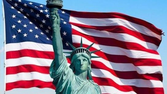 The Statue of Liberty standing in front of a large, billowing American flag against a clear sky.