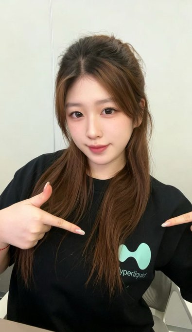 A young woman with long brown hair wearing a black Hyperliquid t-shirt, pointing at the logo with both hands.