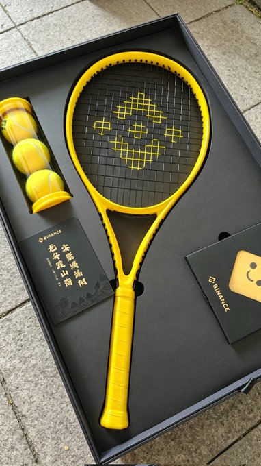 The image features a tennis racket, three tennis balls, and some books or cards placed together. The tennis racket appears to be yellow with black text, while the tennis balls are also yellow. There is an accompanying book or card that has Chinese characters on it. The scene suggests that someone might have purchased these items for their tennis practice or hobby, possibly as a gift or for personal use.