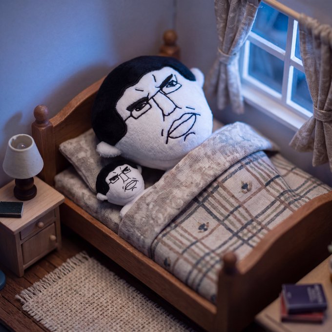 A diorama of a small bedroom with a wooden bed where two plush toys with an angry, drawn-on face are resting under a plaid blanket.
